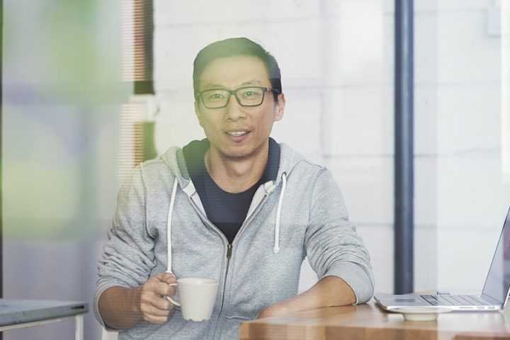 Asian man wearing glasses drinking coffee with arm on table with laptop